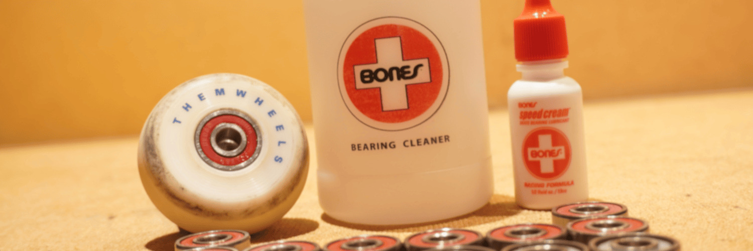 A close-up of a Them Goods skating wheel, Bones Bearing Cleaning Unit, Bones Speed Cream, and Bones bearings scattered around on the back of a picture frame which houses a YuGiOh poster on the front side that's not visible.