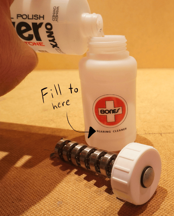 A hand pouring an acetone bottle into a Bones Cleaning Unit that’s next to it’s cap which has 8 bearings attached with a spacer separating each. Overlaid is the hand drawn text “Fill to here” with an arrow pointing at the Bearing Cleaner label on the bottle
