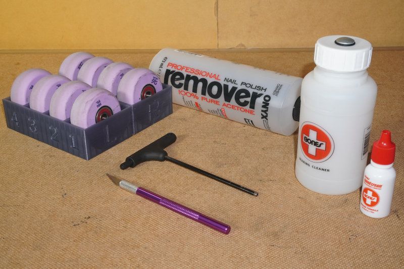 Materials for cleaning bearings scattered around a work area which include 8 purple wheels with bearings in a container, a T-tool handle wrench, a hobby knife, a bottle of nail polish remover, Bones Bearing Cleaning Unit, and Bones Speed Cream