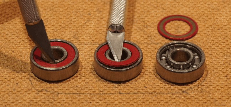 Three bearings lined up. Going left to right, first one has a hobby knife wedged into the shield gap, second has the knife partially popping the shield out, and third has the bearings exposed with the removed shield placed beside it.